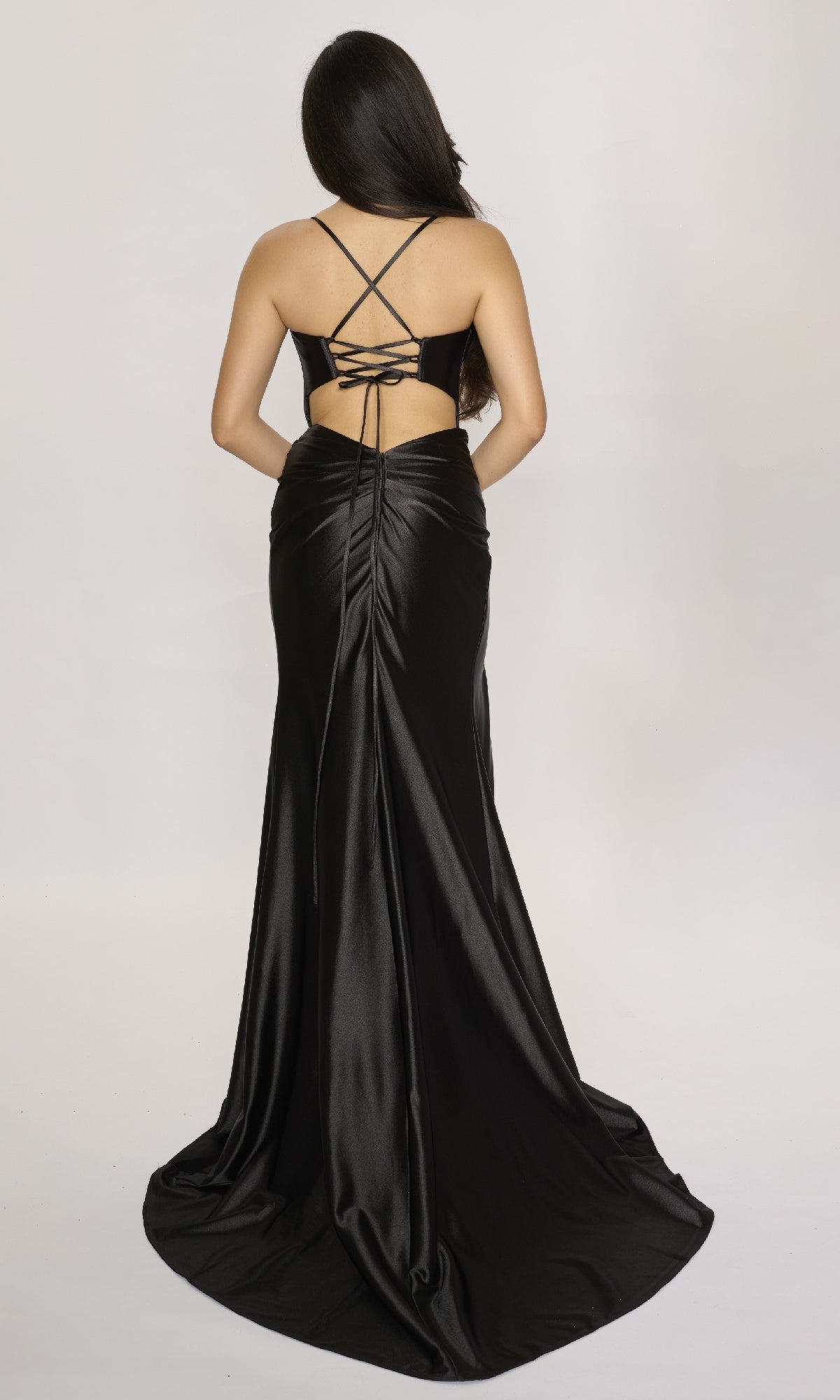 A woman wearing the Long Prom Dress: Nina Canacci 4435—a black satin gown with crisscross straps, open back, and wrap-style leg slit—stands against a plain light background, facing away from the camera.